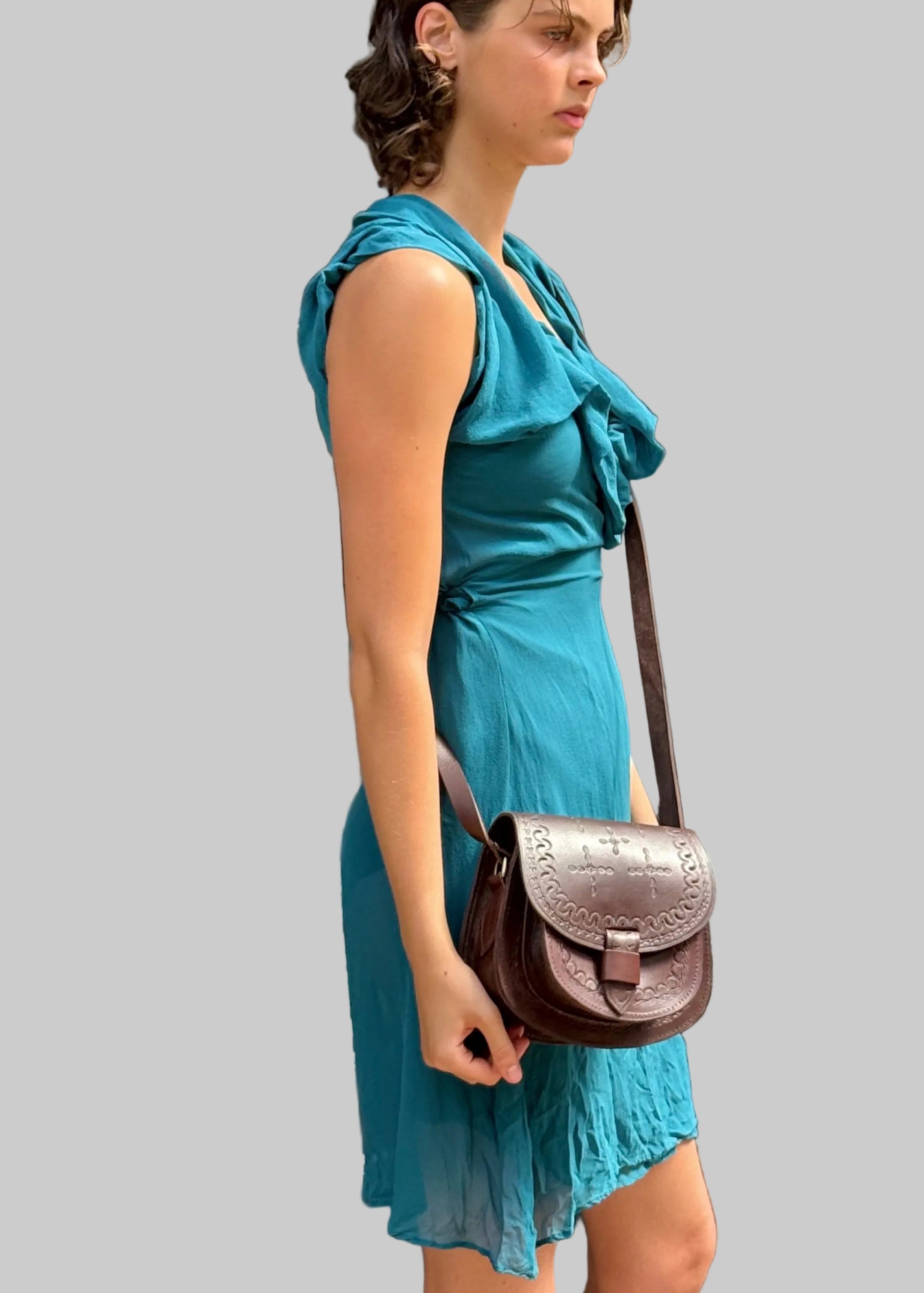 A woman wearing a teal sleeveless dress stands in front of a clothing store window, carrying a brown leather crossbody bag with a flap and long strap.