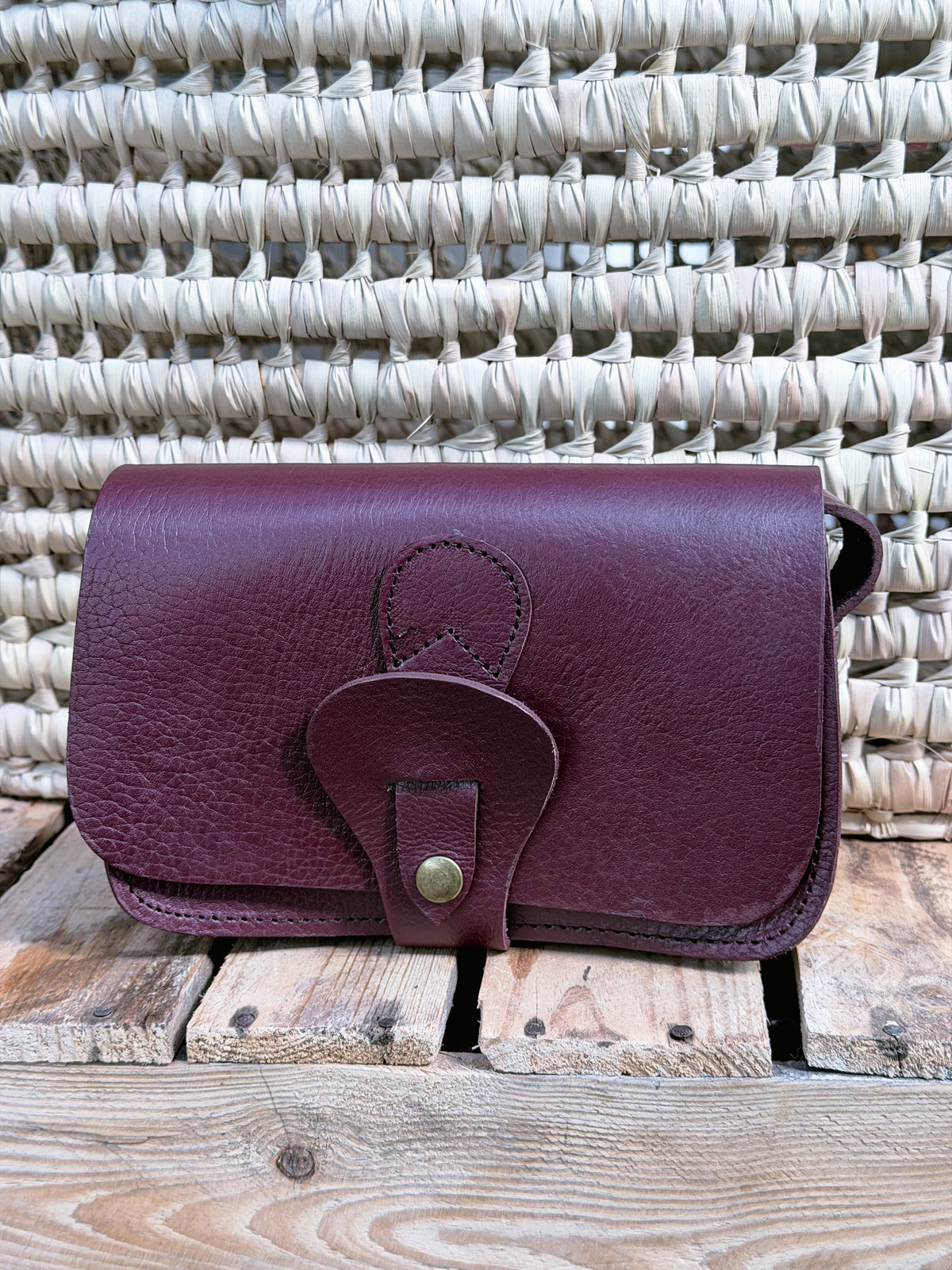 A burgundy leather wallet or card holder with a snap-button strap closure, displayed on a rustic wooden surface in front of a woven straw background.